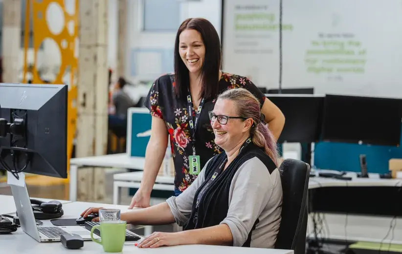 Two female professionals smiling and collaborating at a computer in an open-plan office, illustrating Now NZ's scalable solutions that support business growth and expansion.