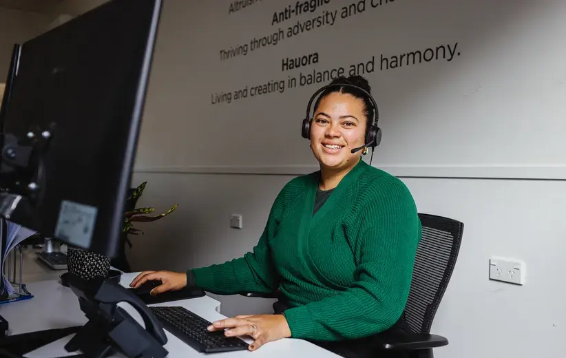 Female customer service representative wearing a headset and smiling while working at a computer in an office, representing Now NZ's award-winning customer service for the health and wellness industry.