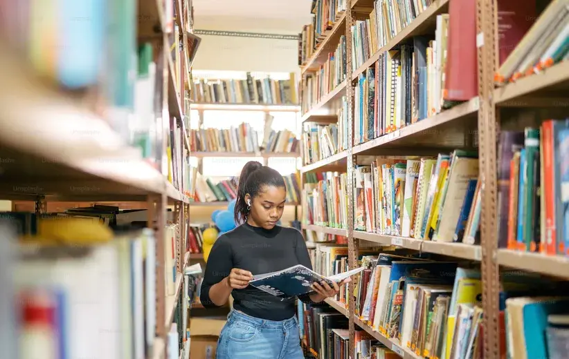Female student reading a book in a library aisle, representing Now NZ's technology solutions for the education and training sector to enhance teaching and simplify administration.