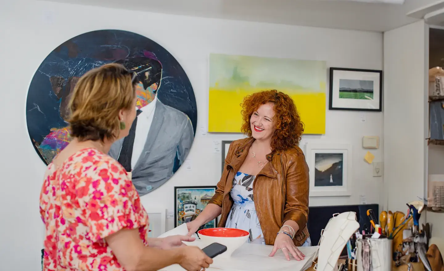 Two women smiling and conversing in an art gallery, one holding a smartphone and the other standing behind a counter with a bowl