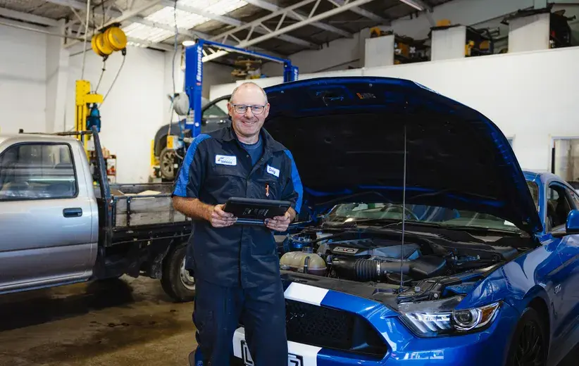 Smiling auto mechanic holding a tablet in a garage next to a car with its hood open, representing Now NZ's telco services tailored for retail and trade businesses to enhance connectivity and opportunities.